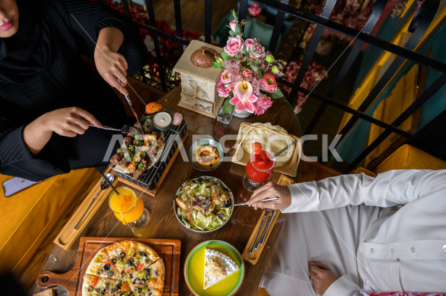 صورة من أعلى لمائدة طعام خاصة بزوجين عربيين خليجيين سعوديين ، يجلسان على مائدة الطعام و يتناولان الطعام ، قضاء اجواء رائعة  في المطعم ، تشكيلة متنوعة من أصناف الطعام و الماكولات الشهية على الطاولة ، جلسات فريدة و مميزة ، مطاعم و كافيهات في السعودية