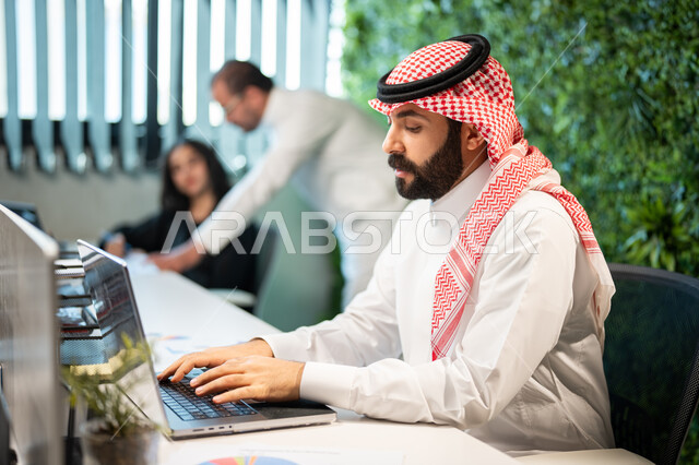 صورة جانبية لموظف عربي سعودي خليجي يرتدي الشماغ و الثوب السعودي التقليدي يقوم بإنجاز مهام العمل اليومية، التواصل مع العملاء من خلال الحاسوب المحمول، تحقيق أهداف الشركة،  التفاهم و التعاون بين الموظفين, بيئة العمل السعودي.
