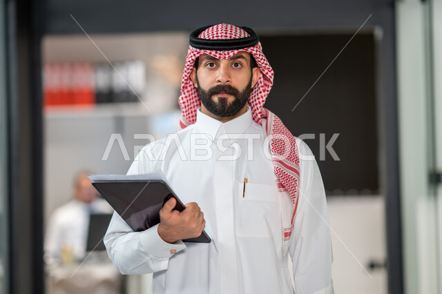 رجل اعمال داخل مقر الشركة، موظف عربي خليجي سعودي يرتدي الشماغ و الثوب السعودي التقليدي يحمل بيده جهاز التابلت و أوراق العمل ينظر الى الكاميرا, مهن ووظائف سعودية، ادارة مهام العمل اليومية،  ايماءات وجه تدل على السعادة، خلفية مكتب.