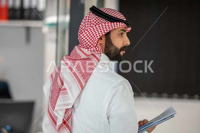 صورة من الخلف لرجل اعمال داخل مقر الشركة، موظف عربي خليجي سعودي يرتدي الشماغ و الثوب السعودي التقليدي يحمل بيده جهاز التابلت و أوراق العمل, مهن ووظائف سعودية، ادارة مهام العمل اليومية،  ايماءات وجه تدل على السعادة، خلفية مكتب.