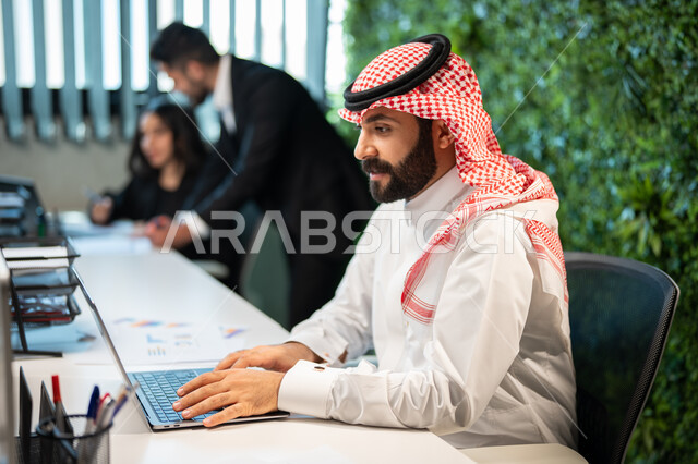 إنجاز مهام العمل اليومية من خلال الحاسوب المحمول، صورة جانبية لموظف عربي سعودي خليجي يرتدي الشماغ و الثوب السعودي التقليدي, التواصل مع العملاء، تحقيق أهداف الشركة،  التفاهم و التعاون بين الموظفين, بيئة العمل السعودي. 