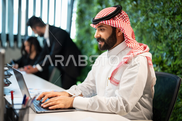 صورة جانبية لموظف عربي سعودي خليجي يرتدي الشماغ و الثوب السعودي التقليدي يقوم بإنجاز مهام العمل اليومية، التواصل مع العملاء من خلال الحاسوب المحمول، تحقيق أهداف الشركة،  التفاهم و التعاون بين الموظفين, بيئة العمل السعودي. 
