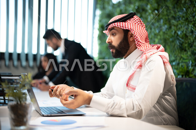 التواصل مع العملاء من خلال مكالمة الفيديو عبر الحاسوب المحمول, صورة جانبية لموظف عربي سعودي خليجي يرتدي الشماغ و الثوب السعودي التقليدي يقوم بإنجاز مهام العمل اليومية، تحقيق أهداف الشركة،  ايماءات وجه و يدين تدل على الاندماج و التركيز, بيئة العمل السعودي.