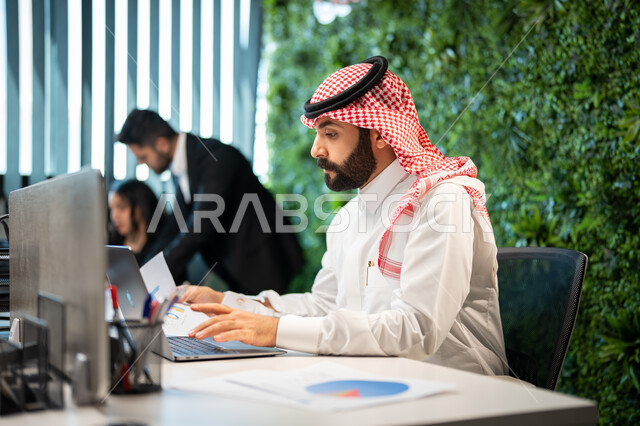 مقارنة النتائج و تدوين الملاحظات, موظف عربي سعودي خليجي يرتدي الشماغ و الثوب السعودي التقليدي يقوم بإنجاز المهام اليومية، دمج الأعمال مع التقنية و التكنولوجيا, تحقيق أهداف الشركة، التفاهم و التعاون بين الموظفين, بيئة العمل السعودي.