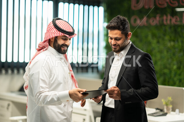 Unity and teamwork to develop work and solve problems, two Saudi Arabian Gulf businessmen inside the company's headquarters discussing work tasks using a tablet, Gulf companies and offices, achieving the company's goals, Saudi professions and jobs, office