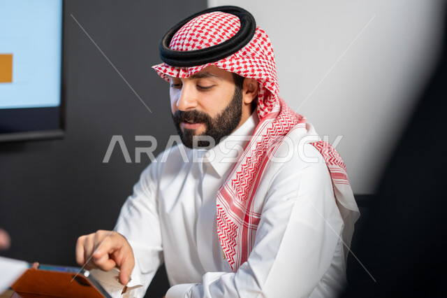 إجراء مكالمات فيديو و الرد على العملاء عبر جهاز التابلت, صورة مقربة لموظف عربي سعودي خليجي يرتدي الشماغ و الثوب السعودي التقليدي يقوم بإنجاز مهام العمل اليومية، تحقيق أهداف الشركة،  ايماءات وجه و يدين تدل على الاندماج و التركيز, بيئة العمل السعودي.