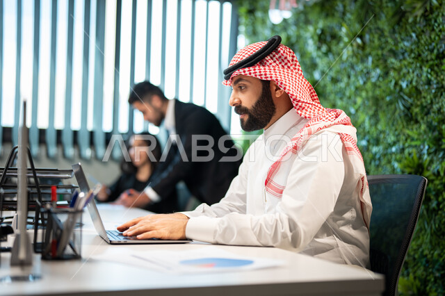 مناقشة وشرح مهام العمل عن بعد، موظف عربي خليجي سعودي يرتدي الشماغ والثوب السعودي التقليدي يقوم بالتواصل مع العملاء من خلال الحاسوب المحمول، إنجاز مهام العمل اليومية، تحقيق أهداف الشركة، بيئة العمل السعودي.