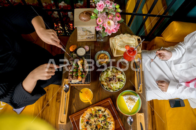 صورة من أعلى لمائدة طعام خاصة بزوجين عربيين خليجيين سعوديين ، يجلسان على مائدة الطعام و يتناولان الطعام ، قضاء اجواء رائعة  في المطعم ، تشكيلة متنوعة من أصناف الطعام و الماكولات الشهية على الطاولة ، جلسات فريدة و مميزة ، مطاعم و كافيهات في السعودية