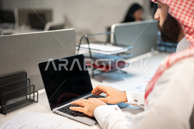 مهن ووظائف سعودية، تحقيق أهداف الشركة، مناقشة وشرح مهام العمل عن بعد، صورة مقربة لموظف عربي خليجي سعودي يرتدي الشماغ والثوب السعودي التقليدي يقوم بإنجاز مهام العمل عبر الحاسوب، بيئة العمل السعودي.