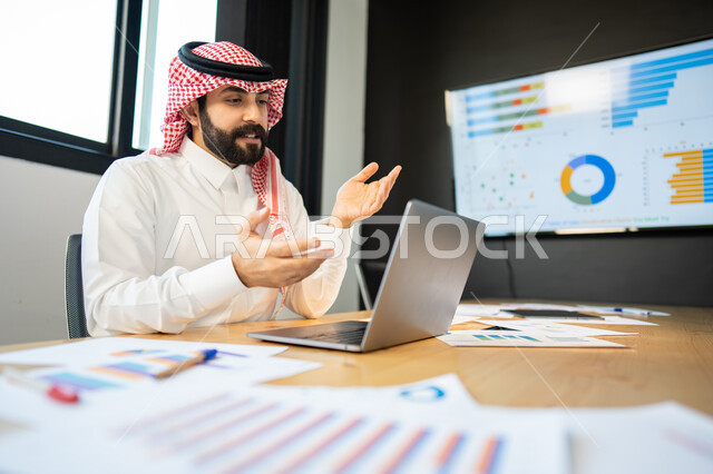 بيانات خسائر وارباح الشركه, شرح و مناقشة مهام العمل عن بعد, ايماءات وجه و يدين تدل على الاندماج و التركيز,  موظف عربي سعودي خليجي يرتدي الزي السعودي التقليدي يقوم بالتواصل مع العملاء بإجراء مكالمة الفيديو عبر الحاسب المحمول, إنجاز مهام العمل اليومية.