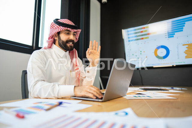 شرح و مناقشة مهام العمل عن بعد, بيانات خسائر وارباح الشركه, ايماءات وجه و يدين تدل على الترحيب و السلام, موظف عربي سعودي خليجي يرتدي الزي السعودي التقليدي يقوم بالتواصل مع العملاء بإجراء مكالمة الفيديو عبر الحاسب المحمول, إنجاز مهام العمل اليومية.