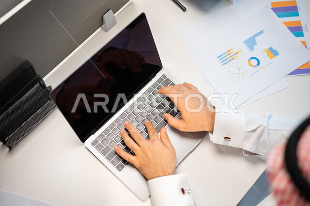 Stock exchange charts, achieving the company's goals, discussing and explaining remote work tasks, a close-up of a Saudi Arabian Gulf employee wearing the traditional Saudi shemagh and dress, completing work tasks via the computer, the Saudi work environm