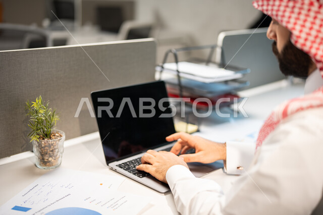 رسوم بيانية للبورصة، تحقيق أهداف الشركة، مناقشة وشرح مهام العمل عن بعد، صورة مقربة لموظف عربي خليجي سعودي يرتدي الشماغ والثوب السعودي التقليدي يقوم بإنجاز مهام العمل عبر الحاسوب، قسم التحليلات والإحصائيات البيانية.
