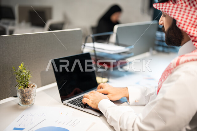 Graphs of the Stock Exchange, achieving the company's goals, discussing and explaining remote work tasks, a close-up of a Saudi Arabian Gulf employee wearing the traditional Saudi shemagh and thobe, completing work tasks via the computer, the analysis and