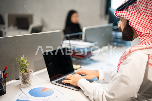 رسوم بيانية للبورصة، تحقيق أهداف الشركة، مناقشة وشرح مهام العمل عن بعد، صورة مقربة لموظف عربي خليجي سعودي يرتدي الشماغ والثوب السعودي التقليدي يقوم بإنجاز مهام العمل عبر الحاسوب، قسم التحليلات والإحصائيات البيانية.