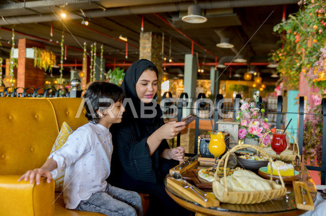 A Saudi Arabian Gulf woman spends wonderful times in the restaurant with her son, holds a mobile phone in her hand and takes a special picture of the dining table in the restaurant, a variety of food items and delicious foods on the table, unique and dist