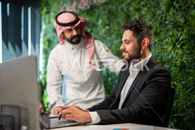 مفهوم الإدارة المتعاونة، موظفان عربيان خليجيان سعوديان يناقشان مهام العمل باستخدام الحاسوب، قسم التحليلات والإحصائيات البيانية، الوحدة والعمل الجماعي لتحقيق أهداف الشركة وحل المشكلات.
