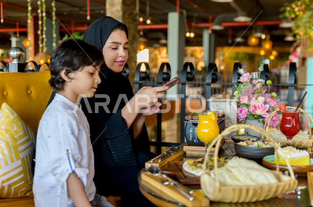 A Saudi Arabian Gulf woman spends wonderful times in the restaurant with her son, holds a mobile phone in her hand and takes a special picture of the dining table in the restaurant, a variety of food items and delicious foods on the table, unique and dist