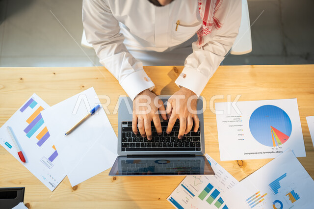 إعداد تقارير عن السوق المالي، متابعة مخطط الرسم البياني للنمو الاقتصادي، صورة علوية عامودية مقربة لموظف يرتدي الزي السعودي ينجز مهام العمل باستخدام الحاسوب، قسم التحليلات والإحصائيات البيانية، بيانات خسائر وارباح الشركة.