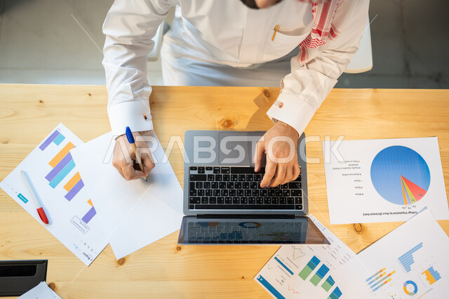 إعداد تقارير عن السوق المالي، متابعة مخطط الرسم البياني للنمو الاقتصادي، صورة علوية عامودية مقربة لموظف يرتدي الزي السعودي ينجز مهام العمل باستخدام الحاسوب، قسم التحليلات والإحصائيات البيانية، بيانات خسائر وارباح الشركة.