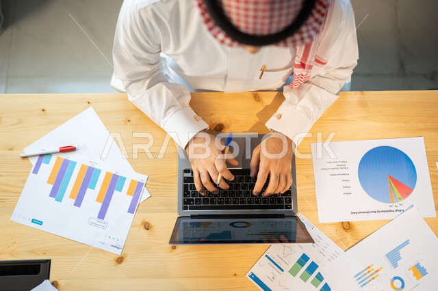 إعداد تقارير عن السوق المالي، متابعة مخطط الرسم البياني للنمو الاقتصادي، صورة علوية عامودية مقربة لموظف يرتدي الزي السعودي ينجز مهام العمل باستخدام الحاسوب، قسم التحليلات والإحصائيات البيانية، بيانات خسائر وارباح الشركة.