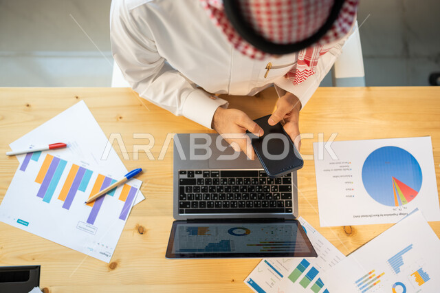 Integration of technology and technology with work, follow-up of the chart of economic growth, an overhead vertical close-up of an employee wearing a Saudi uniform using a mobile phone, the Department of Analytics and Graphic Statistics, the company's pro