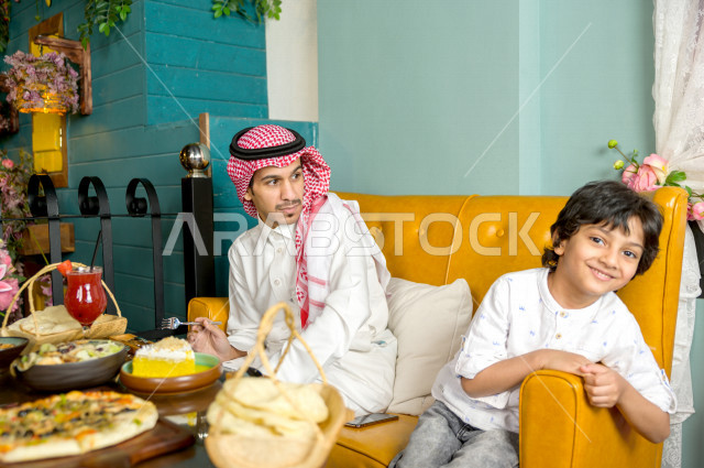 A Saudi Arabian Gulf man is feeding his son in the restaurant with gestures and facial expressions that indicate happiness, spending the holiday with the family, family entertainment atmosphere, unique and distinctive sessions, restaurants and cafes in Sa