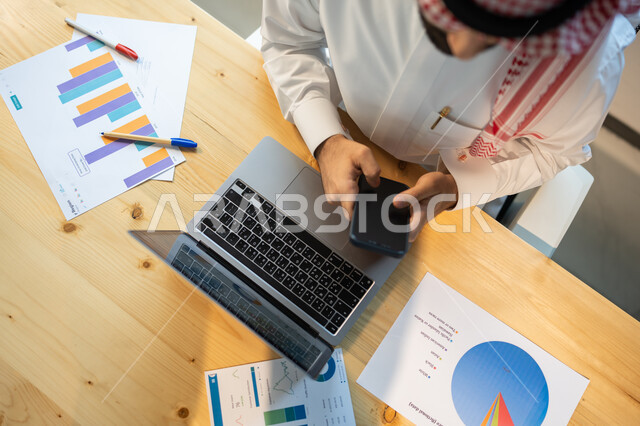 دمج التكنولوجيا والتقنية مع العمل، متابعة مخطط الرسم البياني للنمو الاقتصادي، صورة علوية عامودية مقربة لموظف يرتدي الزي السعودي يستخدم الهاتف المحمول، قسم التحليلات والإحصائيات البيانية، بيانات خسائر وارباح الشركة.