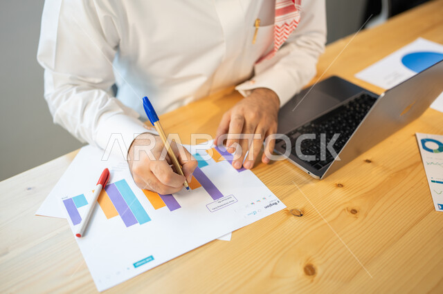 Preparing reports on the financial market, following up the chart of economic growth, a vertical overhead image of an employee wearing a Saudi uniform recording the results, the analysis section and graphic statistics, the company's profit and loss data.