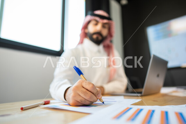 تدوين وتسجيل الملاحظات و النتائج, إعداد تقارير عن السوق المالي، متابعة مخطط الرسم البياني للنمو الاقتصادي، موظف يرتدي الشماغ و الزي السعودي يقوم بإنجاز المهام اليومية ضمن مقر العمل، قسم التحليلات والإحصائيات البيانية، بيانات خسائر وارباح الشركة.
