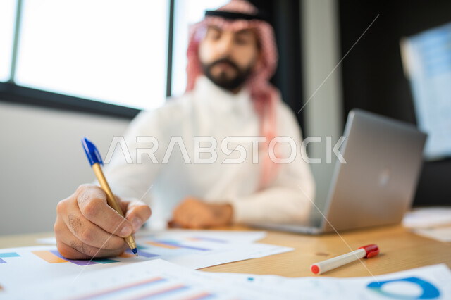 تدوين وتسجيل الملاحظات و النتائج, إعداد تقارير عن السوق المالي، متابعة مخطط الرسم البياني للنمو الاقتصادي، موظف يرتدي الشماغ و الزي السعودي يقوم بإنجاز المهام اليومية ضمن مقر العمل، قسم التحليلات والإحصائيات البيانية، بيانات خسائر وارباح الشركة.