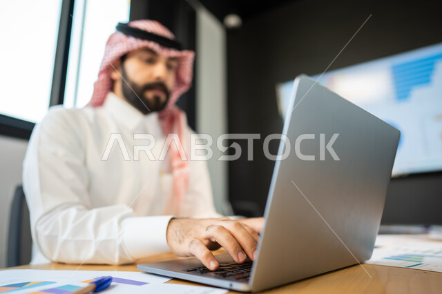 بيانات خسائر وارباح الشركه, ايماءات وجه و يدين تدل على الاندماج و التركيز, موظف عربي سعودي خليجي يرتدي الزي السعودي التقليدي يقوم بإستخدام الحاسب المحمول, دمج التقنية و التكنولوجيا مع الأعمال, إنجاز مهام العمل اليومية.