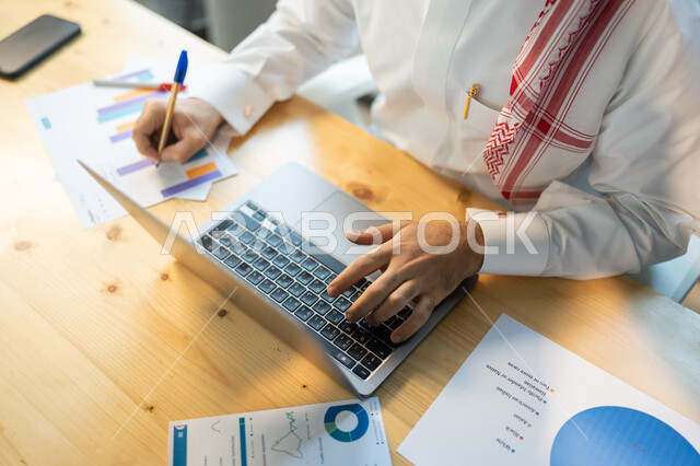 كتابة و إعداد تقارير عن السوق المالي، متابعة مخطط الرسم البياني للنمو الاقتصادي، صورة علوية لموظف يرتدي الزي السعودي يقوم بتدوين النتائج وتسجيل الملاحظات، قسم التحليلات والإحصائيات البيانية، بيانات خسائر وارباح الشركة.