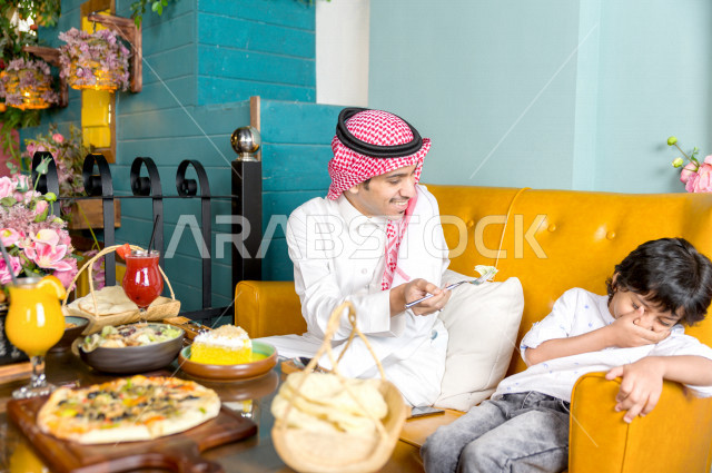 رجل عربي خليجي سعودي يقوم بإطعام ابنه في المطعم بايماءات و تعابير وجه تدل على السعادة ، قضاء العطلة مع العائلة ، أجواء عائلية ترفيهية ، جلسات فريدة و مميزة ، مطاعم و كافيهات في السعودية