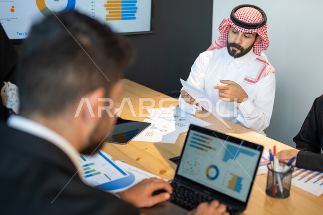 صورة من الخلف لموظف يقوم بدراسة و تحليل النتائج و الرسوم البيانية بإستخدام الحاسب المحمول, قسم البيانات و الإحصائيات, ايماءات تدل على التركيز و الاندماج, دمج التقنية و التكنولوجيا مع البيئة العملية, العمل في روح الفريق.