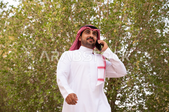 قضاء أوقات ممتعة في الطبيعة الخضراء، الرد على المكالمات الهاتفية عبر الهاتف المحمول، صورة مقربة لرجل عربي خليجي سعودي يقوم بإجراء اتصال هاتفي و التجول في إحدى الحدائق العامة في المملكة العربية السعودية، نزهه في الهواء الطلق, خلفية طبيعة.