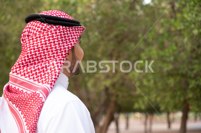 التأمل و قضاء أوقات ممتعة في الطبيعة الخضراء، التجول في إحدى الحدائق العامة في المملكة العربية السعودية، صورة مقربة جانبية لرجل عربي خليجي سعودي يرتدي الشماغ و الثوب السعودي التقليدي, نزهه في الهواء الطلق, خلفية طبيعة.