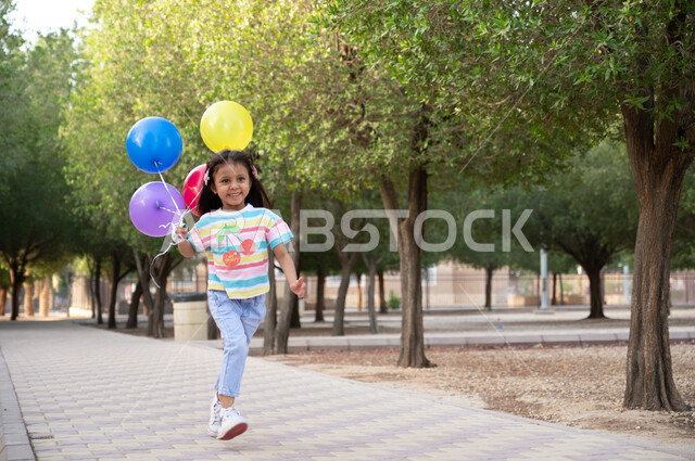 ايماءات وجه تدل على السعادة والسرور بالحصول على بلالين ملونة، طفلة عربية خليجية سعودية تركض في إحدى الحدائق العامة في المملكة العربية السعودية، نزهة في الهواء الطلق والطبيعة الخضراء، أنشطة ترفيهية مسلية للأطفال.