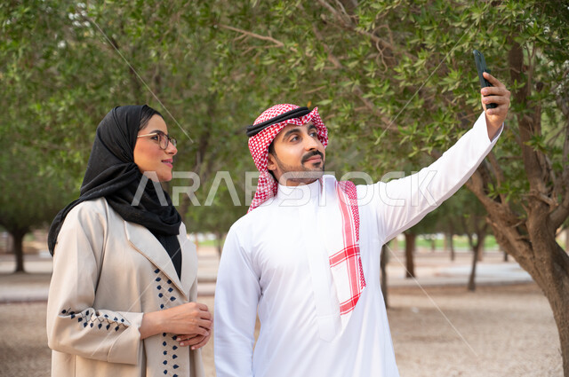 زوجان عربيان خليجيان سعوديان يقومان بإلتقاط صور سيلفي تذكارية بإستخدام الهاتف المحمول, التنزه في حدائق المملكة العربية السعودية, المشي و التجول في الهواء الطلق و الطبيعة الخضراء, أنشطة عائلية ترفيهية. 