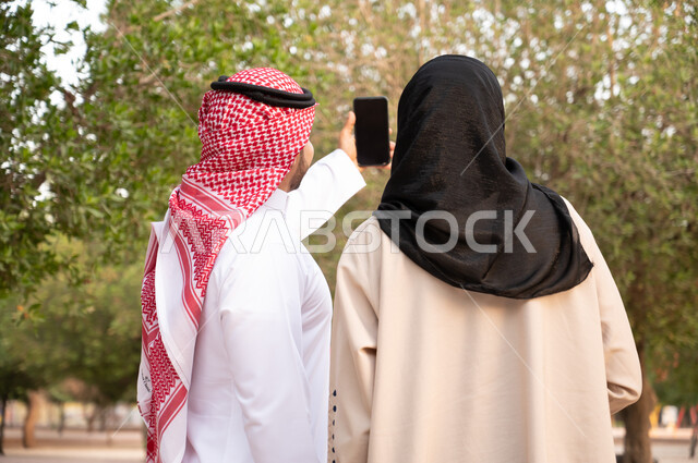 إلتقاط صور سيلفي تذكارية بإستخدام الهاتف المحمول, صورة من الخلف لزوجان عربيان خليجيان سعوديان يقومان بالمشي و التجول في الهواء الطلق و الطبيعة الخضراء, التنزه في إحدى حدائق المملكة العربية السعودية, أنشطة عائلية ترفيهية.