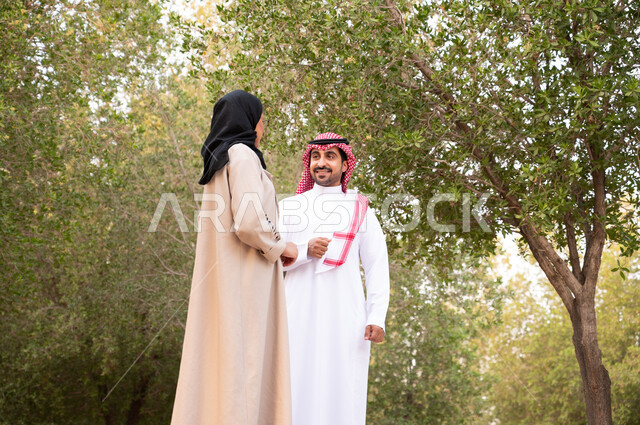 قضاء أوقات ممتعة, تبادل الأحاديث و الأخبار, زوجان عربيان خليجيان سعوديان يقومان بالمشي و التجول في الهواء الطلق و الطبيعة الخضراء, التنزه في إحدى حدائق المملكة العربية السعودية, أنشطة عائلية ترفيهية, خلفية طبيعة.