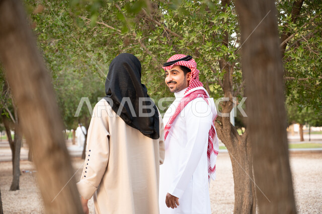 الاستمتاع و قضاء أوقات ممتعة, تبادل الأحاديث و الأخبار, صورة من الخلف لزوجان عربيان خليجيان سعوديان يقومان بالمشي و التجول في الهواء الطلق و الطبيعة الخضراء, التنزه في إحدى حدائق المملكة العربية السعودية, أنشطة عائلية ترفيهية, خلفية طبيعة.