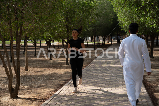 رجل عربي خليجي سعودي يقوم بالمشي في ممر الحديقة, فتاة عربية خليجية سعودية تقوم برياضة الجري للحفاظ على الرشاقة، الصحة واللياقة البدنية و الجسدية، نشاط بدني في الهواء الطلق، التنزه في إحدى الحدائق العامة في المملكة العربية السعودية.