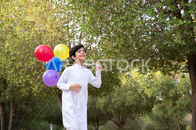 طفل عربي خليجي سعودي يرتدي الثوب السعودي التقليدي يركض في إحدى الحدائق العامة في المملكة العربية السعودية، ايماءات وجه تدل على السعادة والسرور بالحصول على بلالين ملونة، نزهة في الهواء الطلق والطبيعة الخضراء، أنشطة ترفيهية مسلية للأطفال.