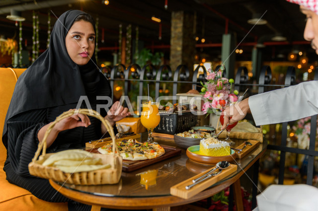 زوجان عربيان خليجيان سعوديان يجلسان على مائدة الطعام و يتناولان الطعام في المطعم ، قضاء اجواء رائعة  في المطعم ، تشكيلة متنوعة من أصناف الطعام و الماكولات الشهية على الطاولة ، جلسات فريدة و مميزة ، مطاعم و كافيهات في السعودية