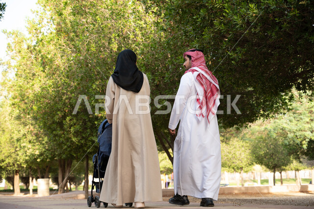 تبادل الأحاديث و الأخبار, صورة من الخلف لزوجان عربيان خليجيان سعوديان يقومان بالمشي برفقة ابنهما الرضيع في الهواء الطلق و الطبيعة الخضراء, التنزه في إحدى حدائق المملكة العربية السعودية, سيدة تدفع بيدها عربة أطفال, أنشطة عائلية ترفيهية, خلفية طبيعة.