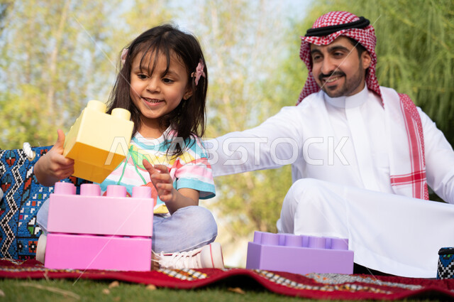 رجل عربي خليجي سعودي يجلس على الأرض بجوار ابنته, اللعب في المكعبات الملونة, ايماءات وجه تدل على السعادة والسرور في أجواء عائلية سعيدة، الحدائق العامة في المملكة العربية السعودية، نزهة في الهواء الطلق والطبيعة الخضراء، أنشطة ترفيهية مسلية للأطفال.