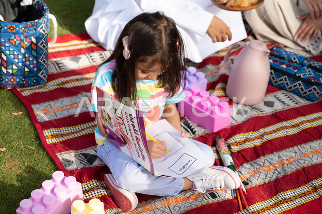 دفاتر و أقلام الرسم و التلوين, أنشطة تعليمية ممتعة للأطفال, فتاة عربية خليجية سعودية تجلس على الأرض و تقوم بالرسم و تلوين الرسومات, ايماءات وجه تدل على السعادة والسرور، الحدائق العامة في المملكة العربية السعودية، نزهة في الهواء الطلق والطبيعة الخضراء.