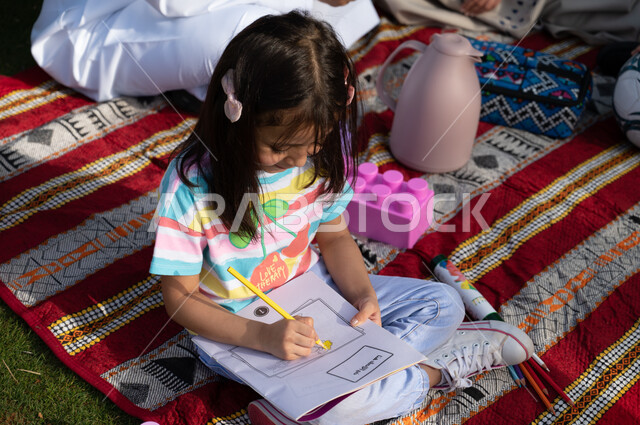 انشطه تعليمية ممتعة للأطفال, دفاتر و أقلام الرسم و التلوين, فتاة عربية خليجية سعودية تجلس على الأرض و تقوم بالرسم و تلوين الرسومات, ايماءات وجه تدل على السعادة والسرور، الحدائق العامة في المملكة العربية السعودية، نزهة في الهواء الطلق والطبيعة الخضراء.