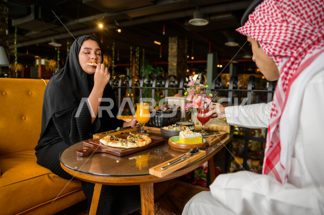 زوجان عربيان خليجيان سعوديان يجلسان على مائدة الطعام و يتناولان الطعام في المطعم ، قضاء اجواء رائعة  في المطعم ، تشكيلة متنوعة من أصناف الطعام و الماكولات الشهية على الطاولة ، جلسات فريدة و مميزة ، مطاعم و كافيهات في السعودية